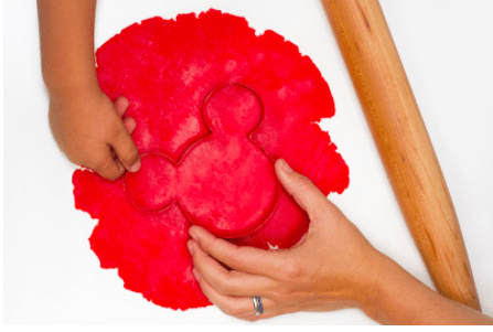 Guests create festive Mickey-shaped holiday cookies from cheerful red dough, with a cookie cutter and rolling pin ready for the magic—just in time for a Disney Parks seasonal celebration.