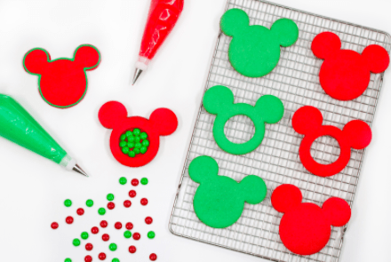 Festive Mickey Mouse-shaped cookies in classic red and green, ready to decorate with colorful icing bags, some featuring a magical Disney twist—hidden red and green candies inside for a sweet holiday surprise.