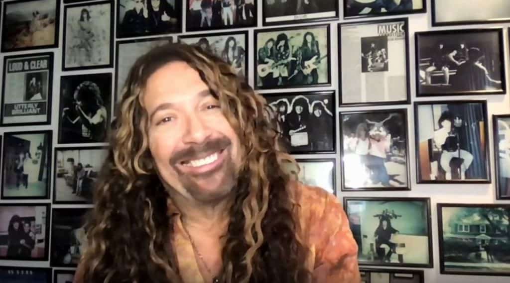 A cheerful man with long, curly hair sits before a wall lined with framed photos of legendary bands and musical moments, capturing the vibe of the voice behind beloved Disney Parks characters like Brer Rabbit from Splash Mountain.