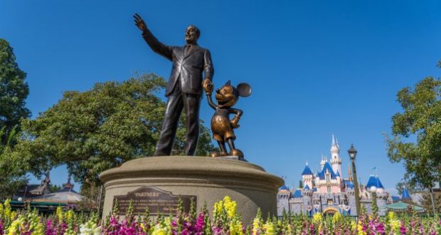 Statue of a man holding hands with a mouse character on a pedestal, surrounded by colorful flowers. A castle with blue and white turrets is in the background under a clear blue sky.