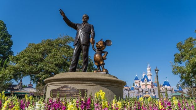 Statue of a man holding hands with a mouse character on a pedestal, surrounded by colorful flowers. A castle with blue and white turrets is in the background under a clear blue sky.