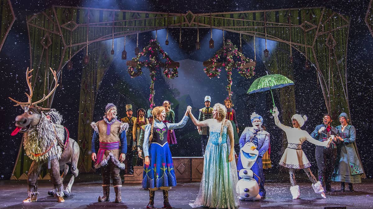 Elsa, Anna, and other Frozen characters light up the stage in vibrant costumes during a snowy musical number, using dazzling props and lively expressions to immerse Disney Cruise Line fans in the magic of an enchanting virtual Frozen Musical experience.