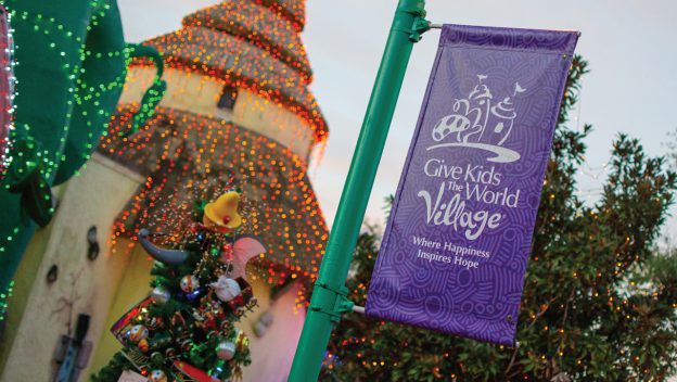 A festive "Give Kids The World Village" banner in bold purple pops beside a beautifully decorated Christmas tree and twinkling lights on the Village’s iconic buildings, creating a scene bursting with whimsical holiday cheer—evoking the enchanting spirit found at top theme park holiday celebrations.