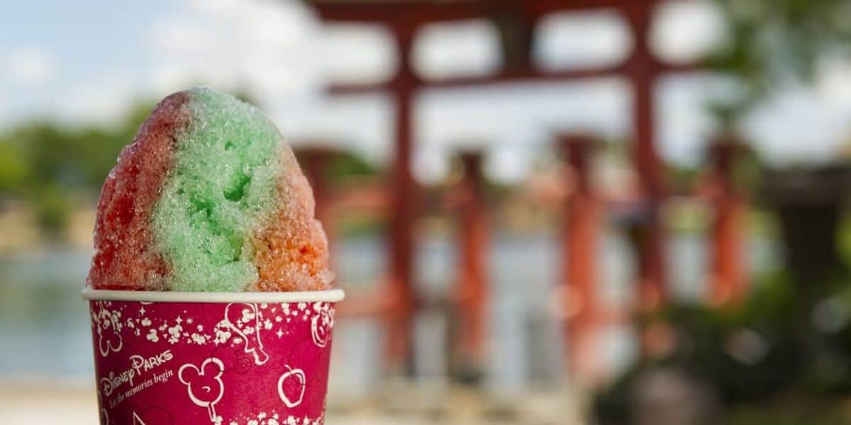A cup of colorful shaved ice with red and green sections in a decorative red cup, set against a blurred background of a traditional Japanese torii gate and a scenic outdoor setting.