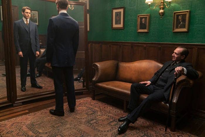 Inside a retro-styled lounge reminiscent of Hollywood Studios, one guest in a sharp suit checks his reflection in the mirror while another relaxes on a brown sofa; their lively discussion buzzes with anticipation for Disney’s highly awaited movie releases.
