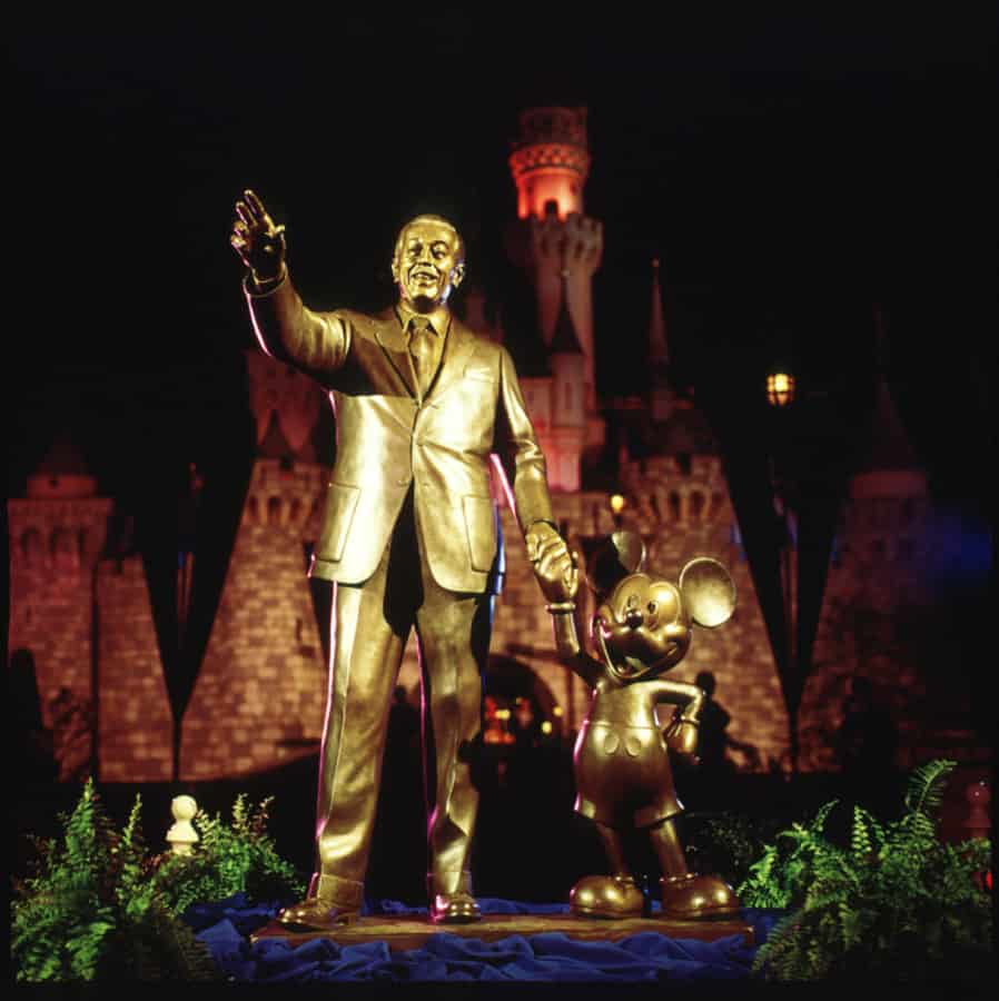 The iconic Partners Statue featuring Walt Disney and Mickey Mouse stands proudly in front of the shimmering Disneyland castle, beautifully illuminated at night and surrounded by lush landscaping—an unforgettable sight for every Disney fan.
