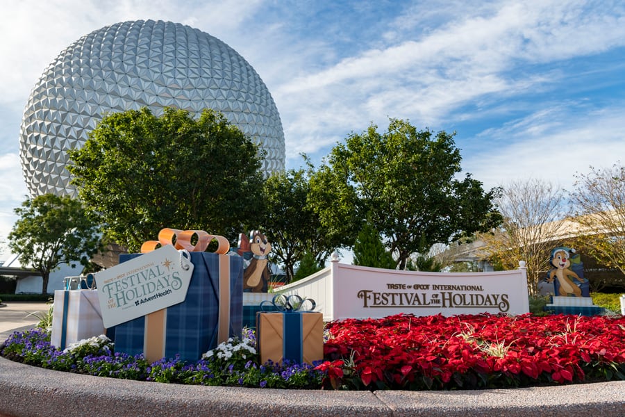Spaceship Earth adorned with Festival of the Holidays signage, colorful gift boxes, and vibrant red poinsettias framing the foreground, sets the festive stage for Disney's EPCOT holiday celebration.