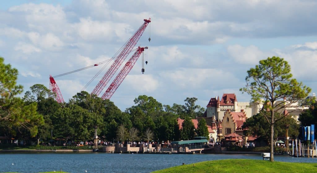 Two towering red cranes rise above the treetops and EPCOT pavilions beside World Showcase Lagoon, set against a partly cloudy sky in this breathtaking aerial view. The scene captures the excitement of ongoing construction for HarmonioUS, echoing the dynamic transformation at Disney’s EPCOT.