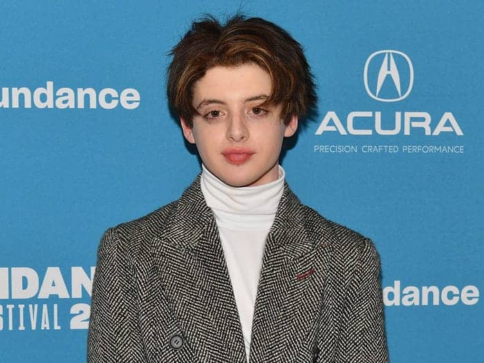 A young celebrity with short brown hair, dressed in a herringbone coat and white turtleneck, strikes a pose at Sundance—capturing the kind of fresh, star-powered look often seen at Disney movie premieres and fan events.