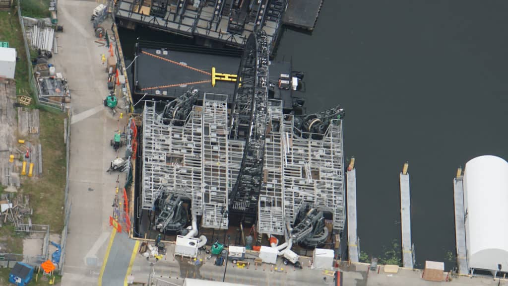 Aerial view of a massive metal bridge being built over a river, surrounded by cranes and construction vehicles, evoking the grand scale and innovation found in Disney park attractions.