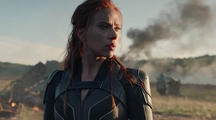 A sharply dressed woman in a sleek black suit stands confidently amid dramatic smoke rising from wreckage, capturing the cinematic excitement and spectacle often seen in Disney's 2021 blockbuster attractions.