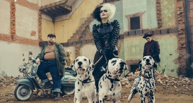 Cruella, dressed in black and surrounded by her three Dalmatians, stands out in front of a backdrop reminiscent of the movie’s edgy London streets; nearby, Jasper and Horace relax with their dog beside a scooter amid artfully distressed scenery—capturing the bold style and cinematic flair Disney brought to life in its 2021 “Cruella” film.