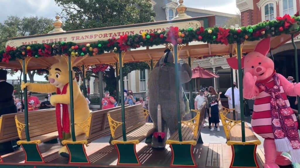 Winnie the Pooh, Eeyore, and Piglet ride atop a brightly decorated trolley adorned with garlands and bows, joining the Magic Kingdom Christmas Cavalcades for a festive holiday celebration.