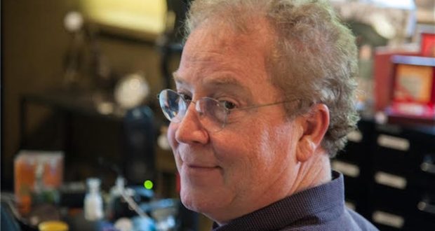 An Imagineer with glasses and curly hair beams at the camera in a creative office space, capturing the inventive energy behind Disney theme park magic.