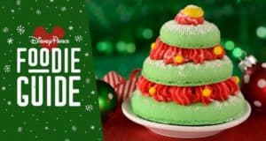 Festive green Christmas tree macaron topped with red frosting and yellow candy, showcased next to the "Disney Parks Foodie Guide"—a magical holiday treat available at Magic Kingdom for Disney fans and foodies to enjoy.