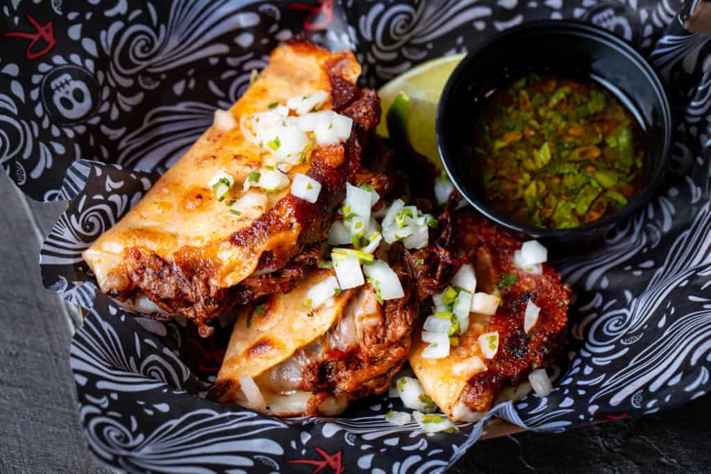 Birria tacos from 4 Rivers Cantina at Disney Springs, topped with onions and cilantro, are served alongside a lime wedge and savory dipping sauce in a black dish—an irresistible treat for theme park foodies enjoying the excitement of Walt Disney World Resort.
