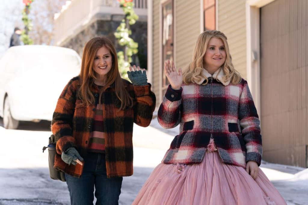 Two women in plaid jackets, one dazzling in a pink gown, wave cheerfully on a snow-dusted, sloping street straight out of Disney+’s Godmothered—capturing the film’s magical winter whimsy as if inviting guests to join in the storybook fun.