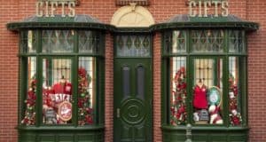 Festively decorated green shopfront with two inviting windows, sparkling with Christmas gifts, cozy sweaters, and whimsical holiday decorations set against a classic brick wall—evoking the enchanting atmosphere you'd find strolling down Main Street during the holidays at a top-tier theme park.