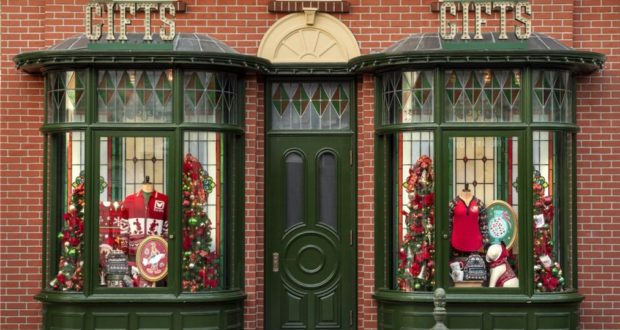 Festively decorated green shopfront with two inviting windows, sparkling with Christmas gifts, cozy sweaters, and whimsical holiday decorations set against a classic brick wall—evoking the enchanting atmosphere you'd find strolling down Main Street during the holidays at a top-tier theme park.