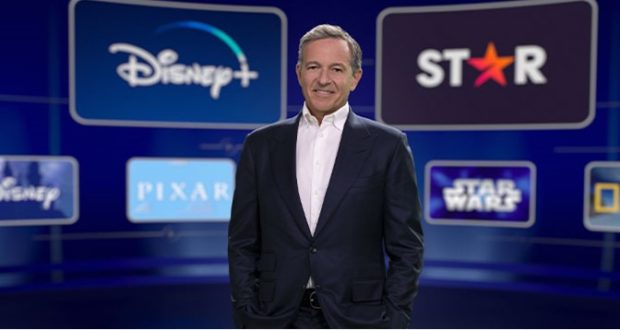 A Disney executive in a suit stands before a colorful backdrop featuring the logos of Disney+, Star, Pixar, Star Wars, and National Geographic, showcasing how Disney’s streaming platforms are shaping entertainment experiences for fans around the world.