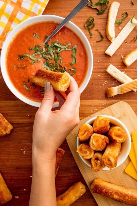 Mickey Mouse dunks a Disney-inspired Grilled Cheese Dipper into rich, creamy tomato soup, surrounded by extra cheesy rolls at a whimsical Disney World table.