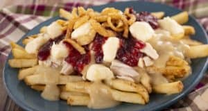 A mouthwatering plate of French fries loaded with savory gravy, melty cheese curds, tender turkey, sweet cranberry sauce, and crispy fried onions on a blue plate—this holiday treat is a culinary highlight at EPCOT Holiday Kitchens. Annual Passholders Discount available.