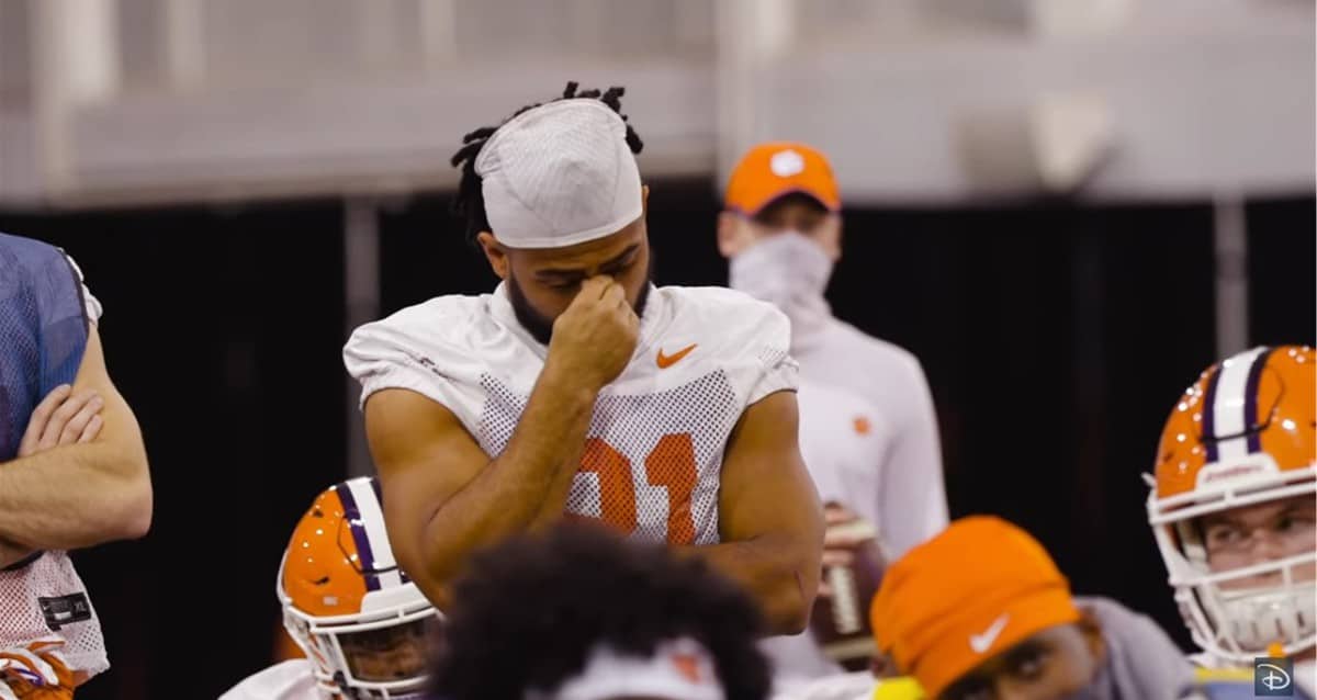 A football player in a white jersey pauses to touch his face during an intense indoor practice with teammates, channeling the determination that earned Clemson Coach Dabo Swinney’s team the 2020 Disney Spirit Award—a testament to Disney’s legacy of celebrating inspiring stories both on and off the field.