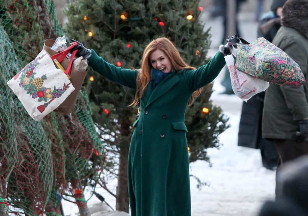 A guest at Disney Springs wearing a festive green coat smiles joyfully while holding shopping bags, surrounded by decorated Christmas trees and falling snow, perfectly capturing the holiday magic and enchantment that feels straight out of a Disney+ movie like Godmothered.
