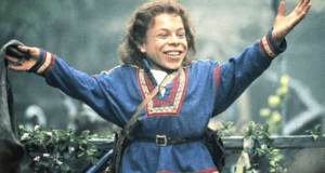 A lively performer dressed in a blue medieval costume greets guests with raised arms amid lush greenery, bringing the adventurous magic of Willow from Disney+ to an outdoor entertainment setting.