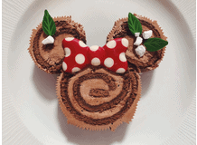 Delicious Chocolate Swiss rolls styled to look like Minnie Mouse, complete with her iconic red polka dot bow and garnished with mint leaves, served on a white plate—this whimsical treat is the ultimate chocolate delight inspired by Minnie’s Yule Log from Disneyland Paris.