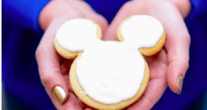 Hands holding a Mickey Mouse-shaped shortbread cookie with a smooth layer of white icing—a whimsical treat that brings the magic of Disneyland’s bakery delights right to your kitchen.