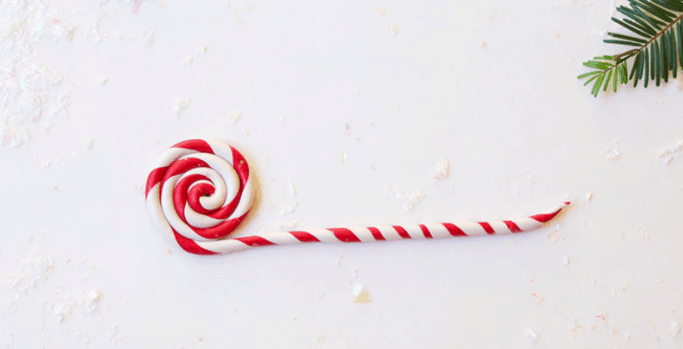 A red and white striped peppermint lollipop sits on a white surface beside a festive green pine branch, evoking Disney Parks holiday treats and channeling the whimsical charm of Mickey Mouse-inspired decorations.