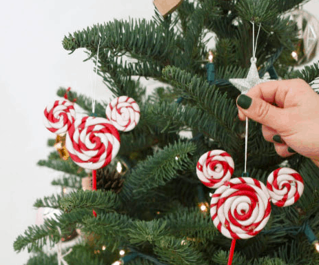 A festive hand adds a sparkling star ornament to a Disney-inspired Christmas tree, decorated with red and white peppermint swirl Mickey Mouse-shaped ornaments—bringing a touch of Disney magic perfect for fans of park-worthy DIY holiday décor.