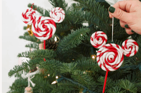 A festive hand places a red and white swirled Peppermint Lollipop ornament, styled after Mickey Mouse, onto a Christmas tree—capturing the magic of Disney holiday decor just like you’d find at the parks.