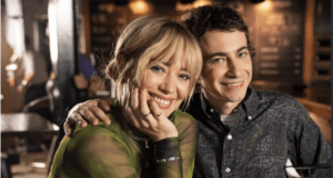 A cheerful duo is pictured indoors, sitting closely as the woman wraps her arm around the man's shoulders, capturing a heartwarming Lizzie McGuire vibe that resonates with Disney+ fans still wishing for the series’ return.