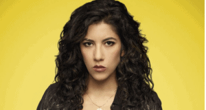 A Disney performer channeling a new animated film heroine, with long curly dark hair and a black jacket, standing in front of a vivid yellow backdrop that echoes the vibrant magic of Encanto meet-and-greets in the park.