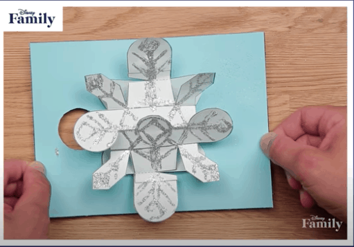 Guests admire a Frozen-inspired pop-up card featuring an intricate blue paper snowflake, set on a rustic wooden table—perfect for sparking creative card-making ideas or commemorating your magical day at the park.