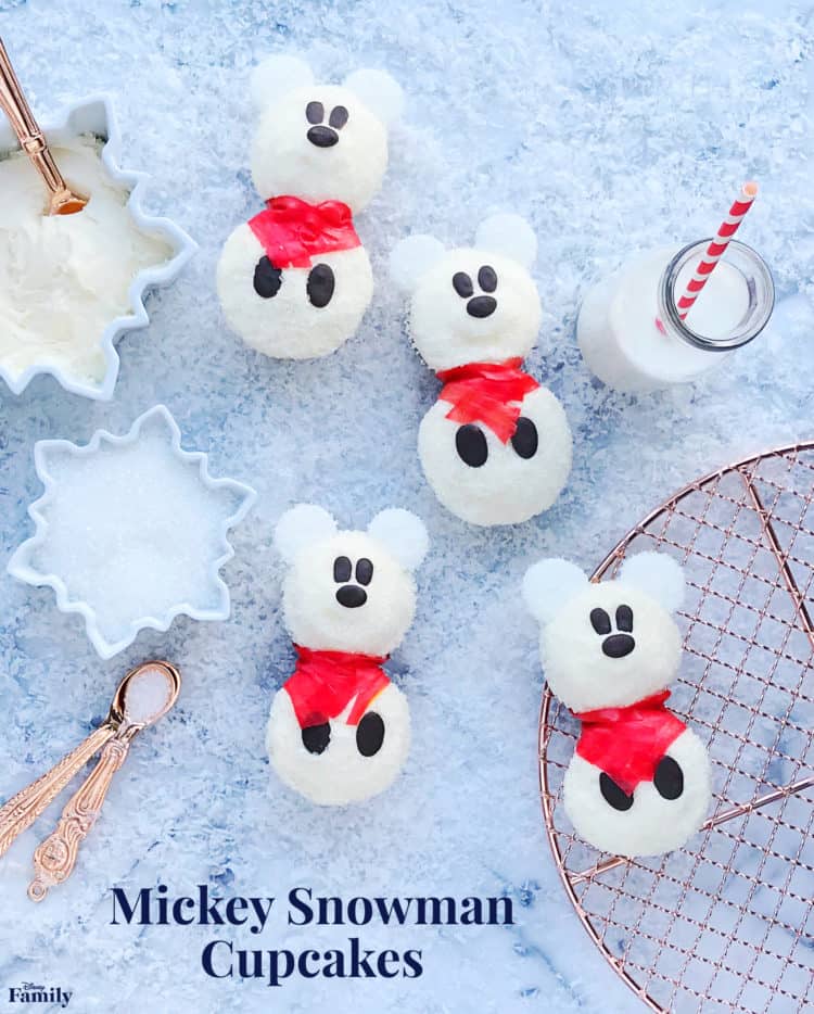 Whimsical Mickey Snowman Cupcakes sporting festive red scarves are displayed on a vibrant blue surface, perfectly paired with a glass of milk and baking tools—bringing Disney magic to your holiday treats.