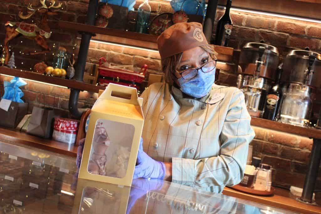 A Ganachery pastry chef in a signature coat and mask presents a yellow pastry box behind the glass counter, celebrating The Ganachery’s 5th Anniversary at Disney Springs.