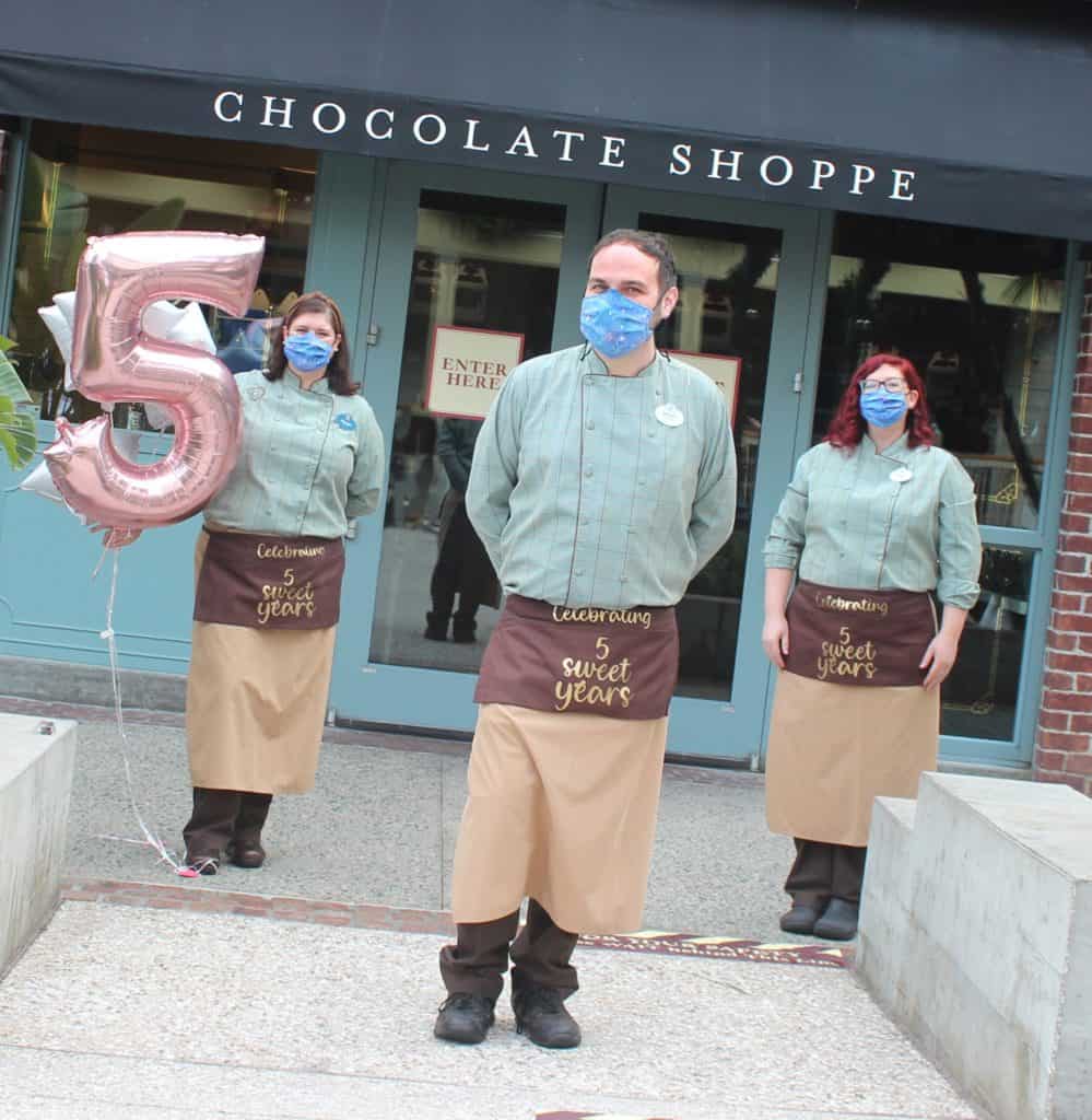 Cast Members in festive aprons and masks stand outside The Ganachery at Disney Springs, proudly displaying a number 5 balloon to mark the shop’s 5th anniversary celebration.