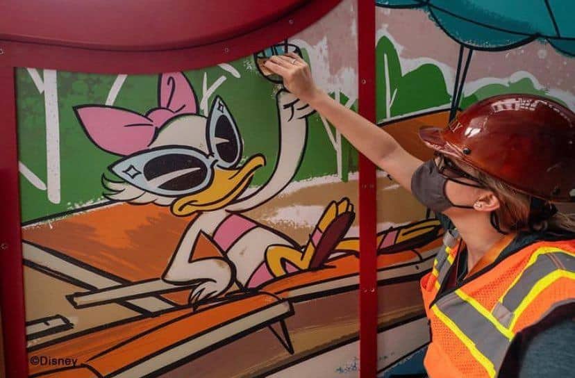Daisy Duck, sporting cool sunglasses, lounges by the iconic Boardwalk Resort pool slide in the lively Keister area as a Cast Member in a hard hat interacts with her vibrant mural.
