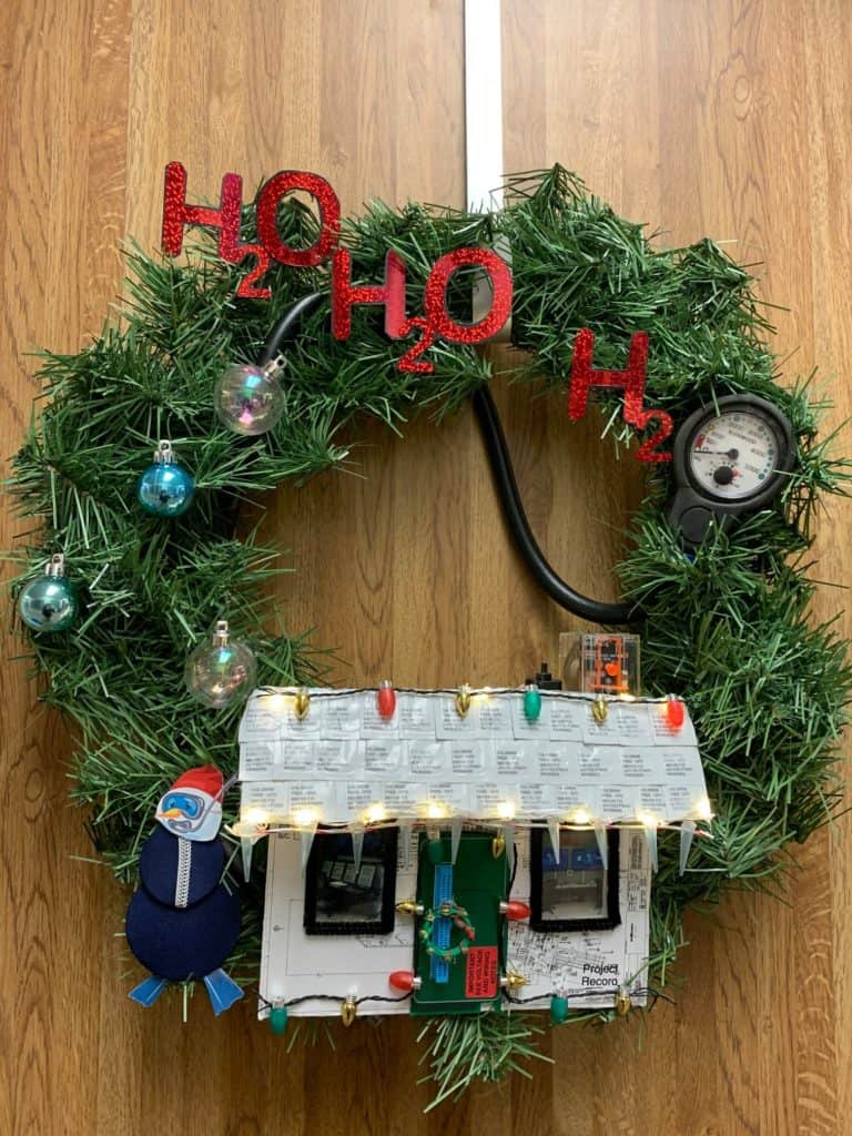 Festive holiday wreath decked out with science lab gadgets, a circuit board, pressure gauge, glowing bulbs, and bold red "H2O" lettering—a clever creation ideal for a Disney Cast Member backstage or as an eye-catching entry in a Disney Parks Wreath-Decorating Contest.