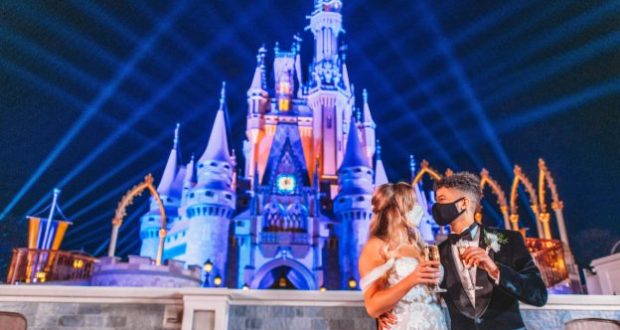 Newlyweds toast in front of Cinderella Castle at Magic Kingdom Park, illuminated by vibrant lights and dramatic beams shooting into the night sky, evoking the enchanting atmosphere of Jordan Fisher and Ellie Woods’ romantic celebration.
