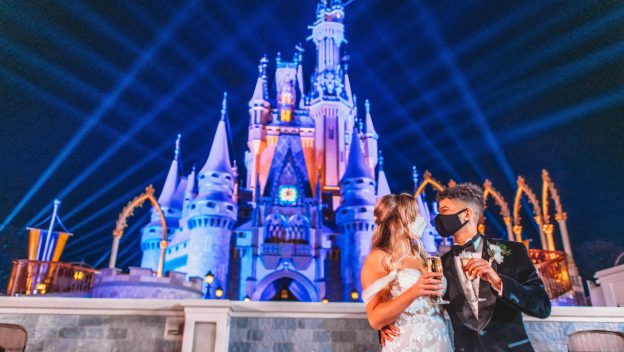 Newlyweds toast in front of Cinderella Castle at Magic Kingdom Park, illuminated by vibrant lights and dramatic beams shooting into the night sky, evoking the enchanting atmosphere of Jordan Fisher and Ellie Woods’ romantic celebration.