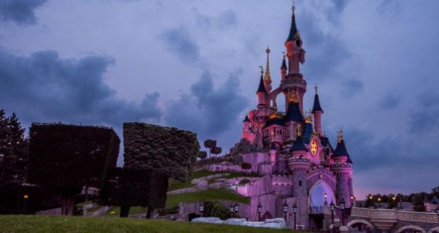 Sleeping Beauty Castle at Disneyland Paris glows with pink lights at dusk, framed by beautifully manicured trees and an enchanting cloudy sky. The iconic centerpiece of the park shines even brighter after its recent refurbishment, capturing all the wonder and magic Disney fans love.
