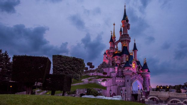 Sleeping Beauty Castle at Disneyland Paris glows with pink lights at dusk, framed by beautifully manicured trees and an enchanting cloudy sky. The iconic centerpiece of the park shines even brighter after its recent refurbishment, capturing all the wonder and magic Disney fans love.