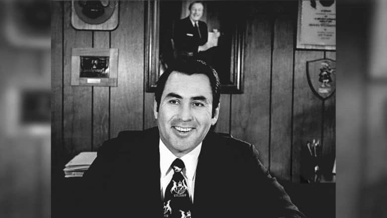 Ron Dominguez, the legendary Disney executive who helped shape the magic of Disneyland, smiles warmly in his suit at his desk. Behind him, a collection of awards and a portrait on wood-paneled walls celebrate his incredible legacy and lasting impact on Walt Disney Attractions, honoring one of Disney’s most beloved figures.
