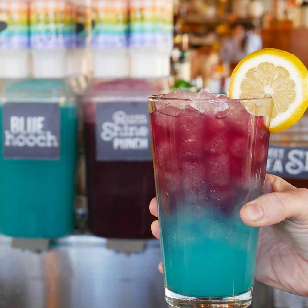The vibrant Purple Lamborghini Moonshine, layered with bold colors, ice, and a lemon slice, is held up for a photo in front of the drink dispensers at Chef Art Smith's Homecomin' at Disney Springs—perfect for guests looking to sip something showstopping during their Walt Disney World adventure.