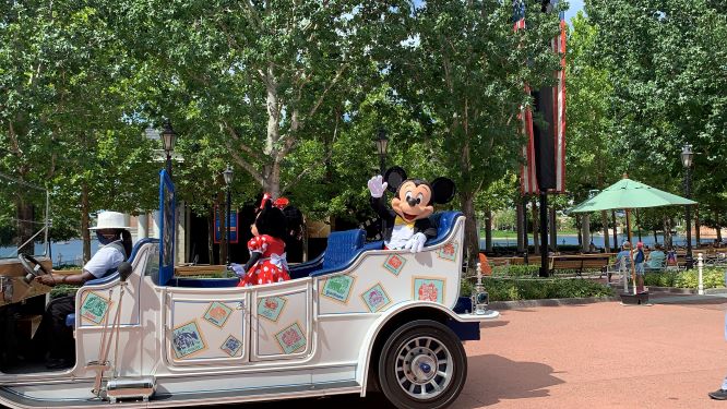 Mickey Mouse and Minnie Mouse wave enthusiastically from a festively decorated white car as they cruise through a sun-drenched, tree-lined path at Walt Disney World, marking the resort’s exciting phased reopening celebration.