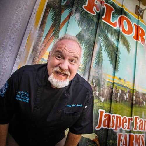A cheerful chef with white hair and a beard stands before a vibrant farm-themed mural at Disney Springs, excited to unveil a new Florida-Inspired Menu in celebration of National Florida Day.